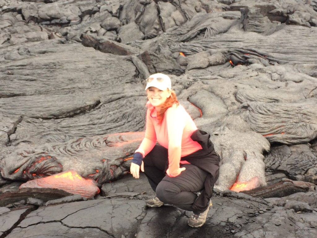 Lainey La Shay crouches near an active lava flow on Kilauea, surrounded by hardened black volcanic rock with glowing orange lava visible beneath the surface.